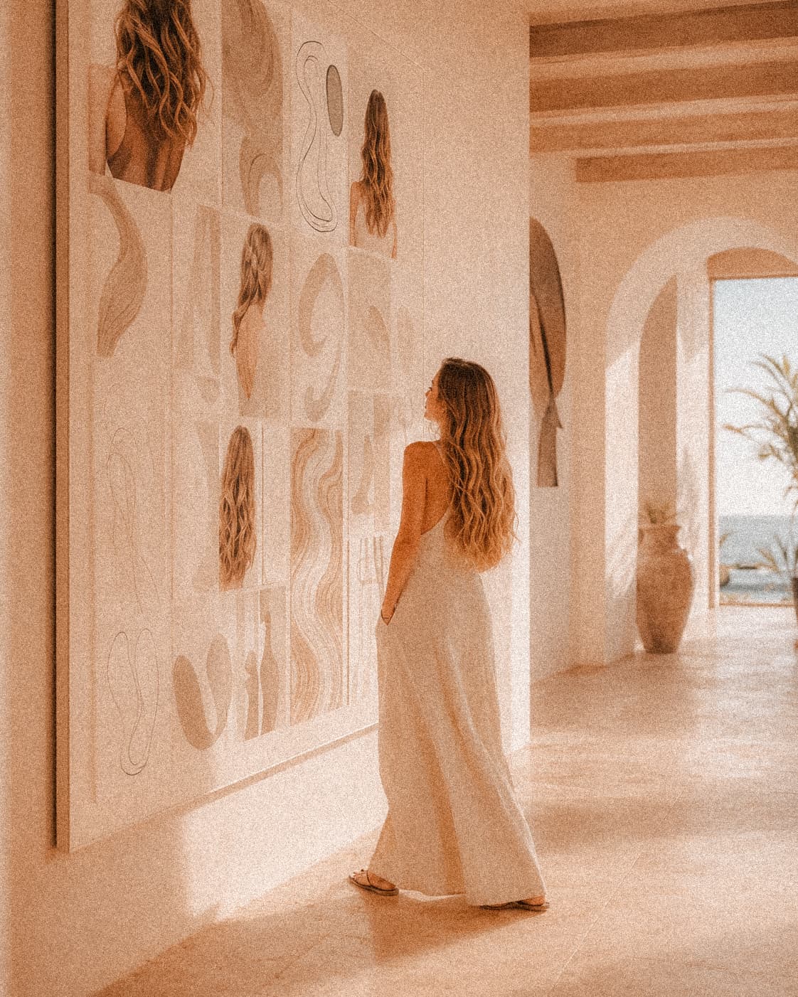 A woman pausing in a sunlit resort hallway to study a floor-to-ceiling wall of hair studies and artwork